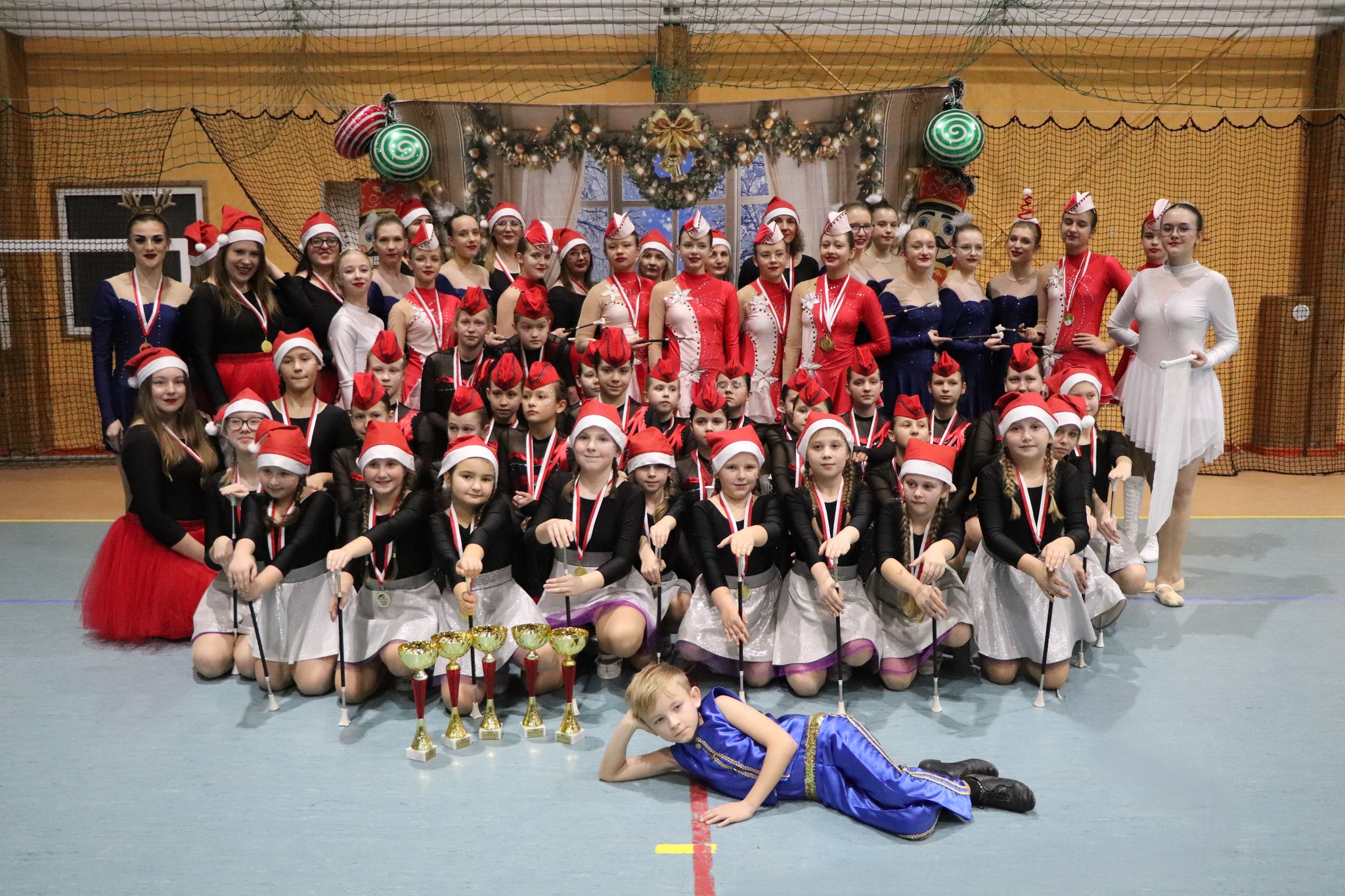 Zdjęcie grupy mażoretek, część dziewczyn ma mikołajowe czapki. Przed grupą stoją ustawione na podłodze puchary. Obok leży chłopiec w niebieskim stroju scenicznym.